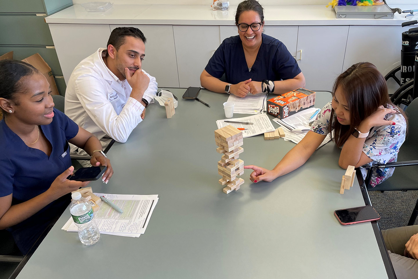 Several fellows sit around large table with papers and a Jenga block tower as one fellow tries to remove a block