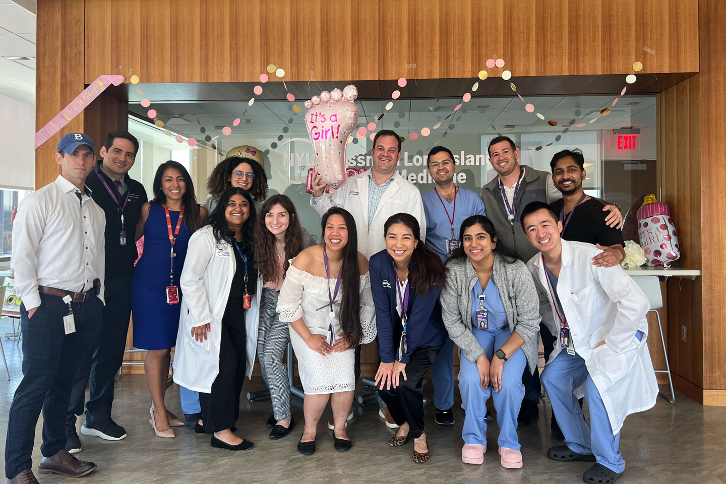 A group of male and femal residents pose for a photo to celebrate a babyshower. One man at the back holds a balloon in the shape of a foot saying “It’s a Girl”.