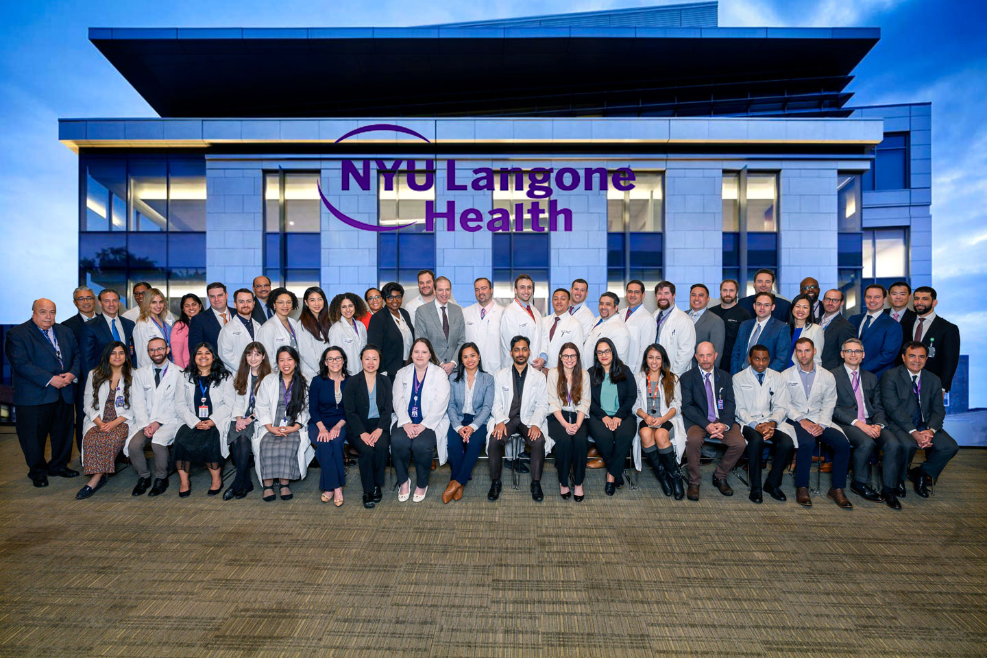 Group photograph of the faculty members and residents at the Department of Surgery. The people stand in two rows in front of a large sign on a building that says NYU Langone Health.