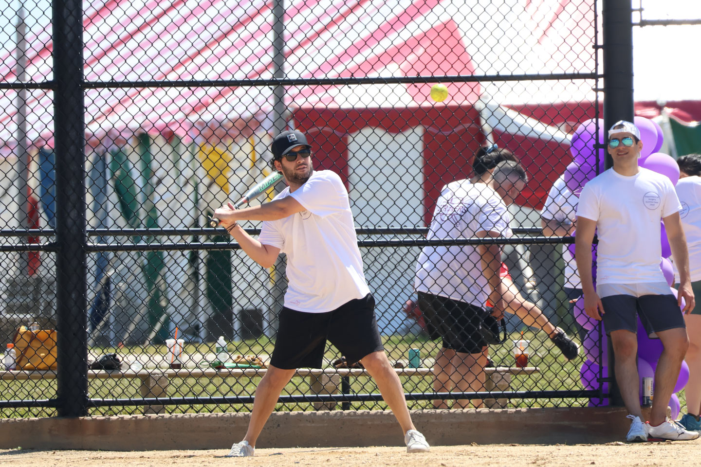 A male resident playing baseball swings at a ball being pitched at him. One man watches, while a group of people behind him are talking.