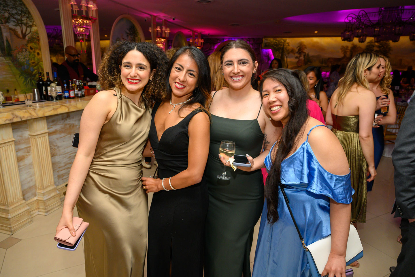 Four female residents in ballgowns of various colors pose for the camera at their graduation ceremony. One is holding a phone and purse, another is holding a phone and glass of wine, another has her handbag over her right shoulder.