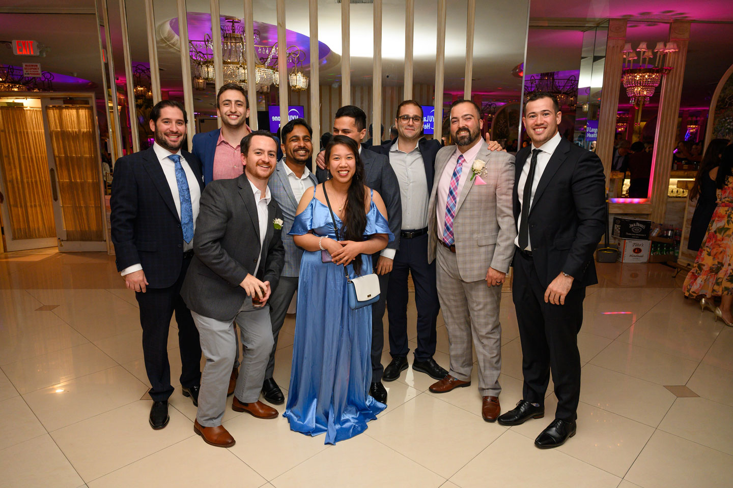 A group of eight men in suits and one female in a ballgown are grouped together to celebrate at their 2024 graduation ceremony.