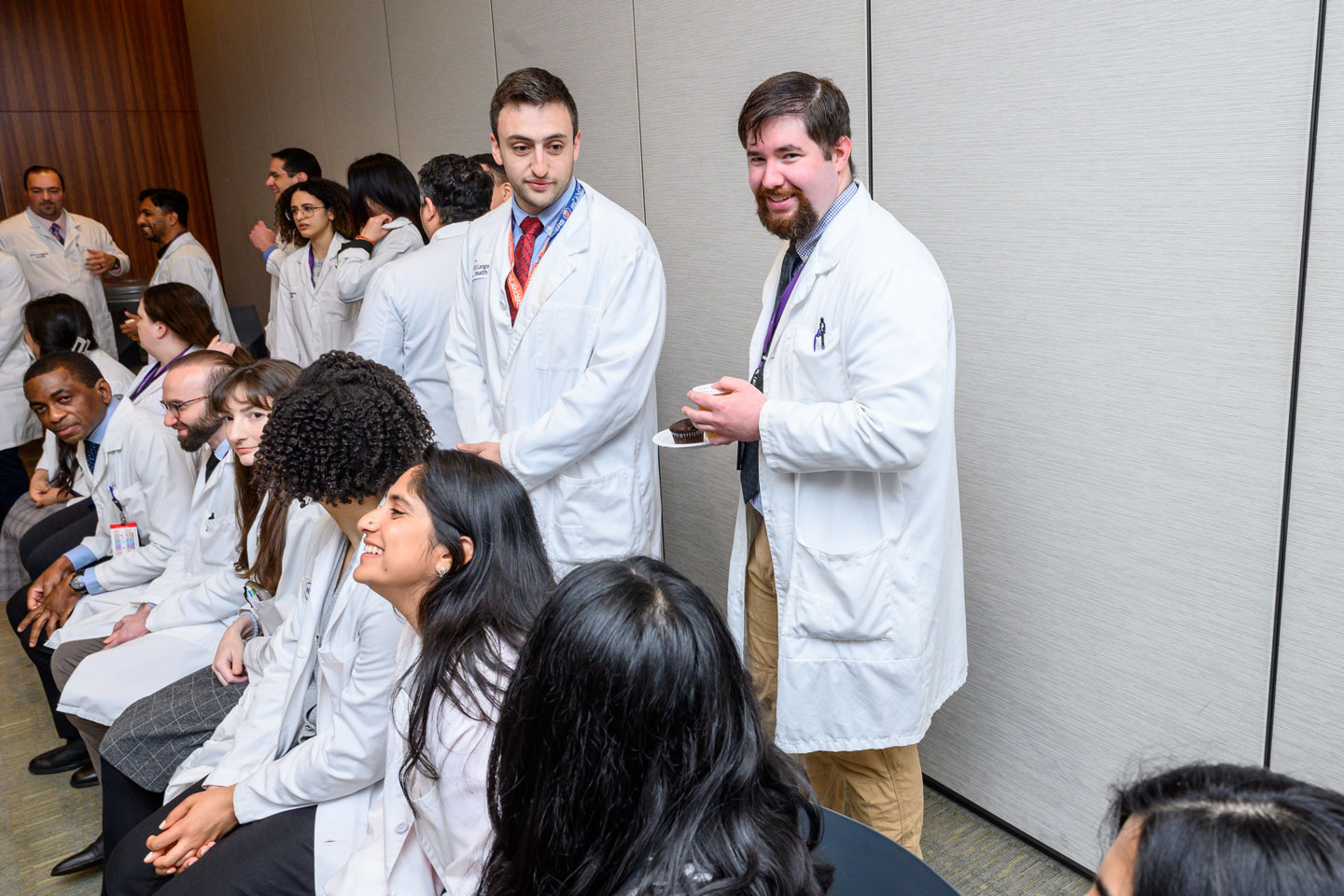 A group of male and female residents wearing labcoats.  A row of residents are sitting down, a group of people behind them are standing up and chatting. 