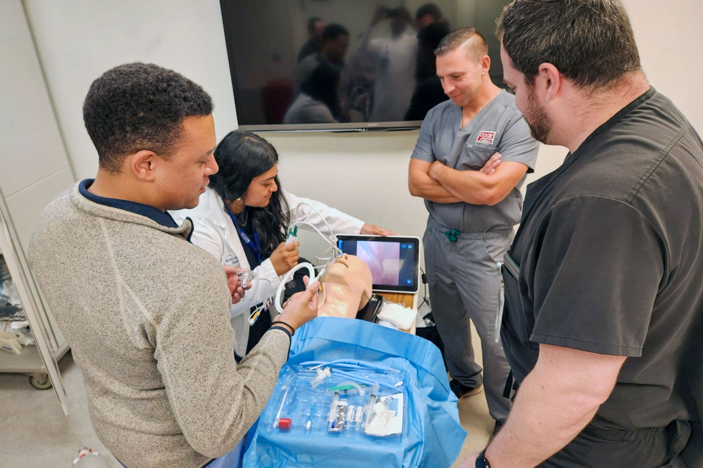 Residents examine a training mannequin with medical equipment while referring to images on a tablet.