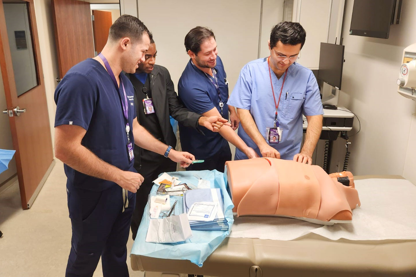 Four medical professionals practice a procedure on a medical training mannequin in a clinical setting.