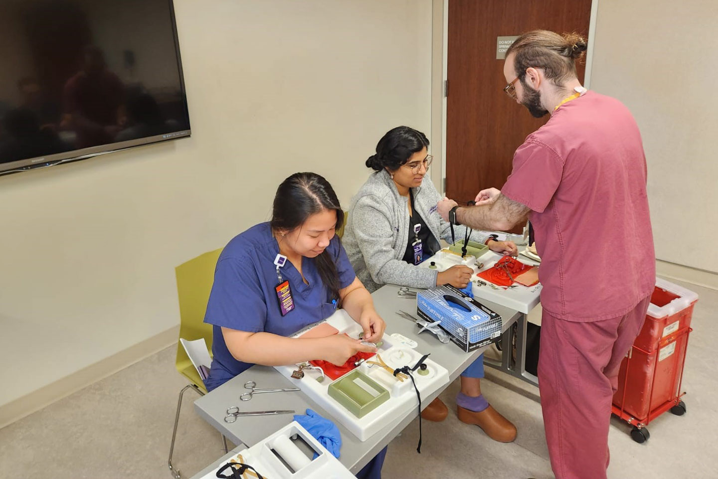 Two seated medical students practice tying knots, while a third individual stands nearby helping.