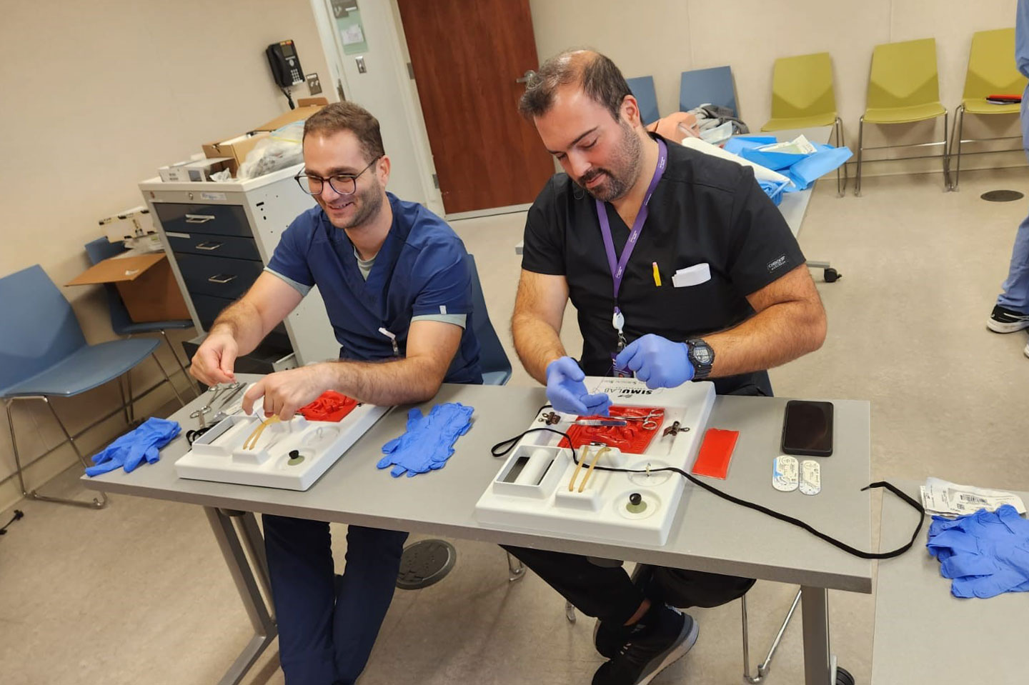 Two seated medical students practice knot tying on simulation kits.